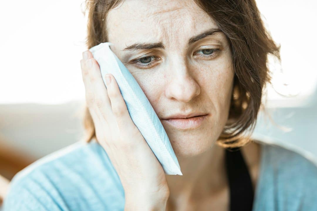 Woman holding a hand to her cheek, easing how long lip filler swelling lasts with a cool compress